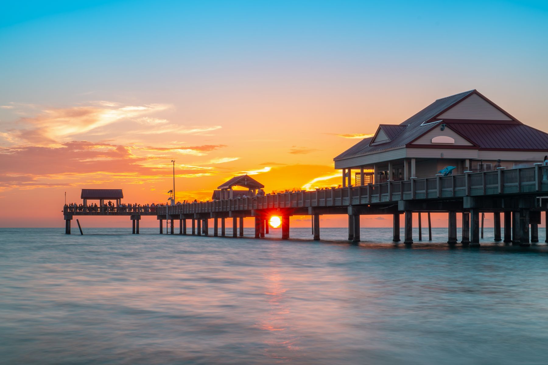 A vibrant sunset over the Gulf of Mexico with a long pier extending into the water under a colorful orange and purple sky.